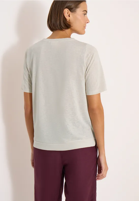 Vrouw van achteren in licht T-shirt en bordeauxrode broek voor een neutrale achtergrond.