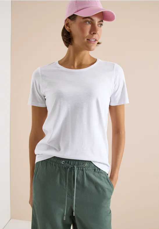 Person trägt weißes T-Shirt, grüne Hose und pinke Cap, steht vor hellem Hintergrund.