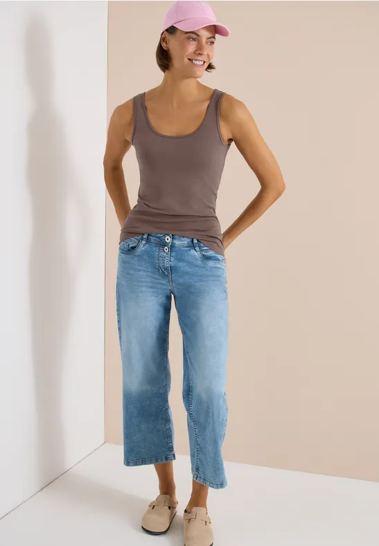 Frau steht in einer Zimmerecke, trägt ärmelloses Top, weite cropped Jeans und Loafers.