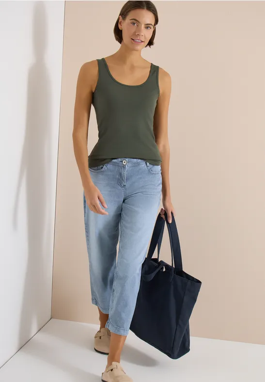 Frau in olivgrünem Tanktop, weiten Jeans und mit dunkelblauer Tasche vor hellem Hintergrund.