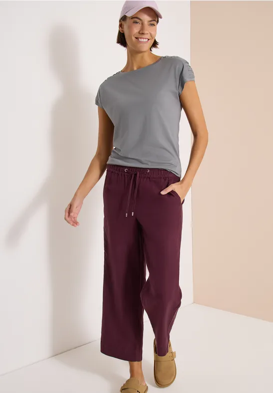 Person steht in einem zweifarbigen Eck, trägt graues T-Shirt, bordeauxrote Hose und Hausschuhe.