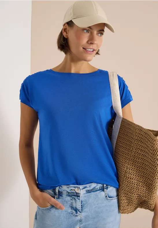 Person trägt blaues T-Shirt, Jeans, weiße Kappe und Strohtasche über der Schulter.