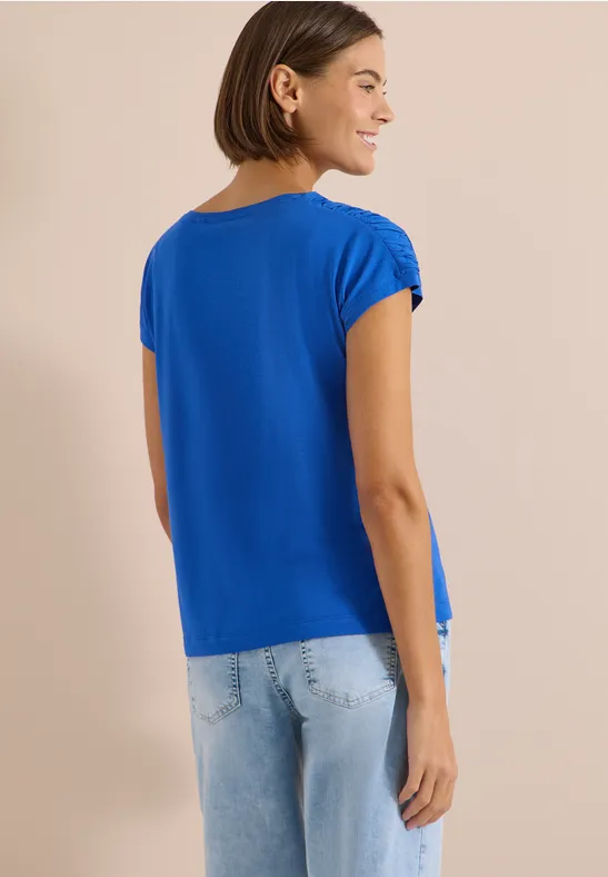 Frau von hinten in blauem T-Shirt und hellen Jeans vor schlichtem, beigem Hintergrund.
