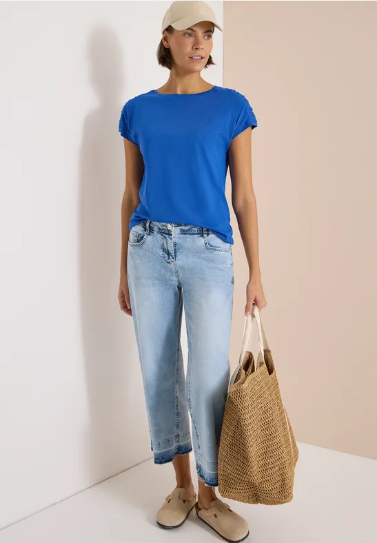 Frau steht in blauem T-Shirt, hellen Jeans, Sneakern und trägt eine große geflochtene Tasche.