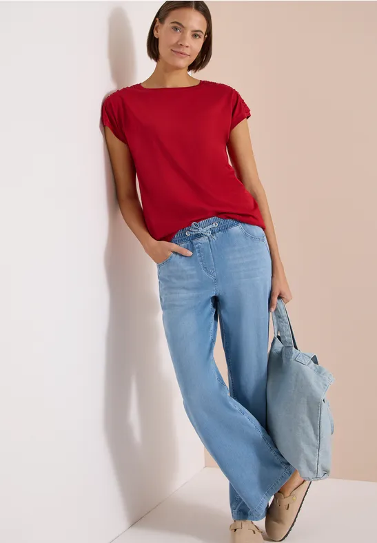 Frau lehnt an weißer Wand, trägt rotes Shirt, weite hellblaue Jeans und hellblaue Tasche.