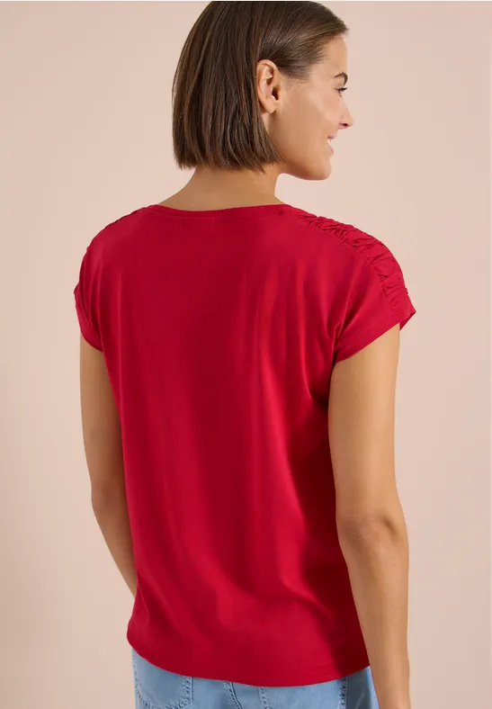Frau mit kurzen braunen Haaren trägt ein rotes kurzärmeliges Shirt, Rückenansicht vor neutralem Hintergrund.