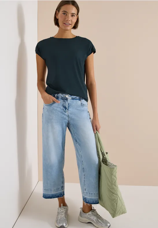 Person trägt schwarzes T-Shirt, weite helle Jeans und hält eine große grüne Stofftasche.