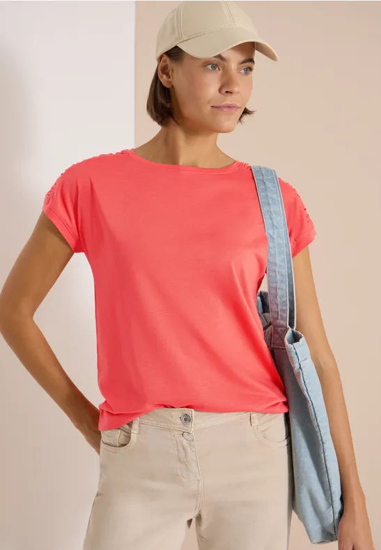 Frau in rotem T-Shirt, beiger Hose und Cap, trägt eine Umhängetasche.