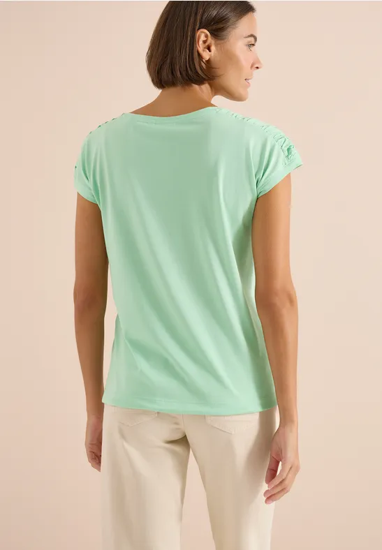 Achteraanzicht van een vrouw in mintgroen T-shirt en lichtbeige broek voor een neutrale achtergrond.