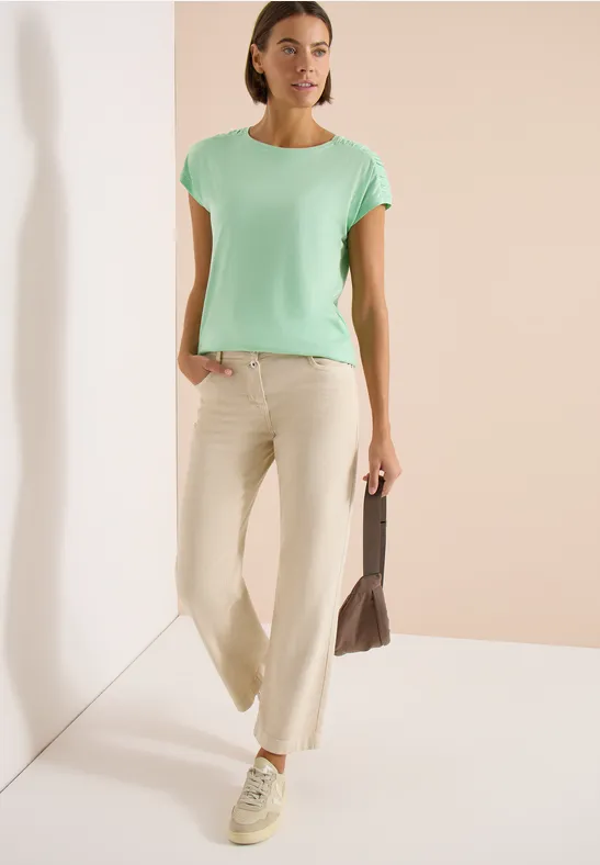 Een vrouw draagt een mintgroen T-shirt, een wijde beige broek en een tas, staand in een lichte ruimte.