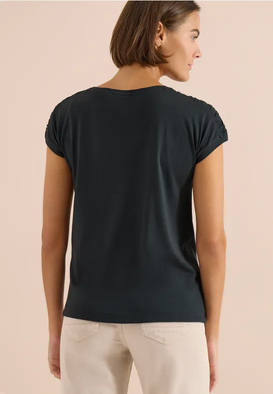 Vrouw van achteren in een donkerblauw t-shirt en lichte broek tegen een neutrale achtergrond.