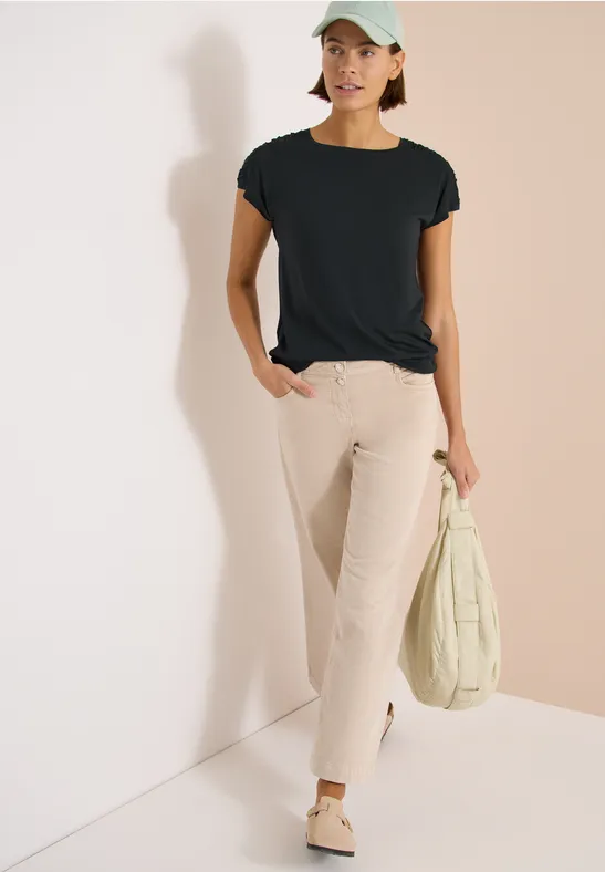 Vrouw leunt tegen lichte muur, draagt zwart shirt, wijde beige jeans, lichte schoenen en houdt een gewatteerde tas vast.