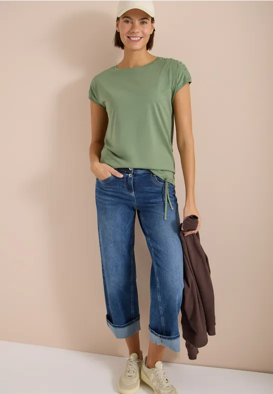 Frau trägt grünes T-Shirt, lässige blaue Jeans, weiße Sneaker, hält braune Jacke in der Hand vor hellem Hintergrund.