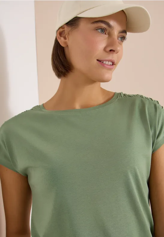 Person trägt grünes T-Shirt und beige Baseballkappe vor hellem Hintergrund.