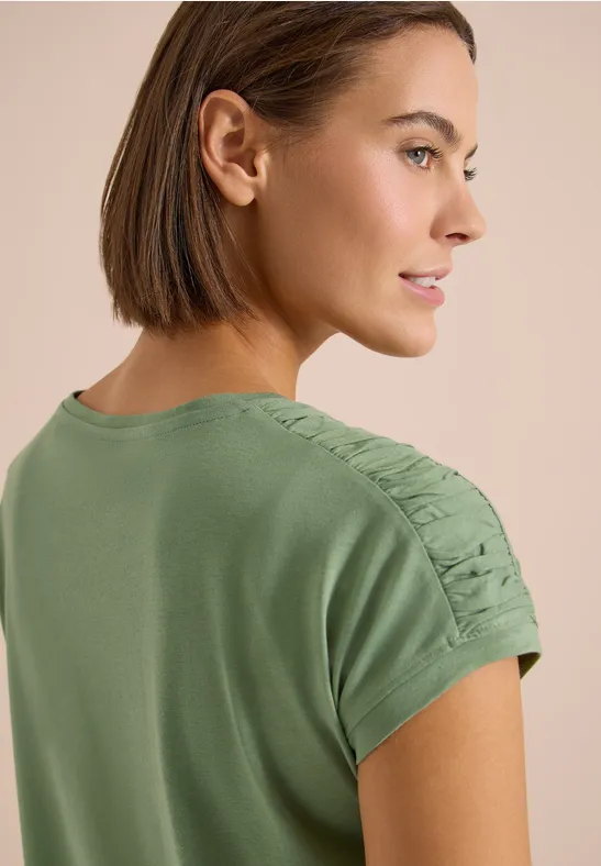 Rückansicht einer Person in khakigrünem Shirt mit kurzer brauner Bob-Frisur und gerafften Ärmeln.