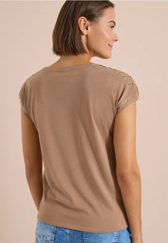 Rückansicht eines Modells in einem beigen T-Shirt mit kurzen Ärmeln und blauer Jeans vor hellem Hintergrund.