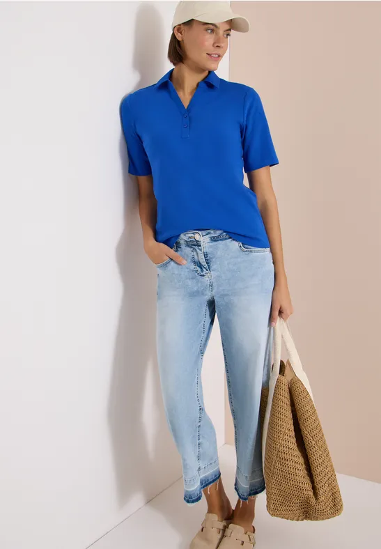 Frau in blauem Poloshirt und hellen Jeans lehnt an Wand und hält eine Korbtasche.