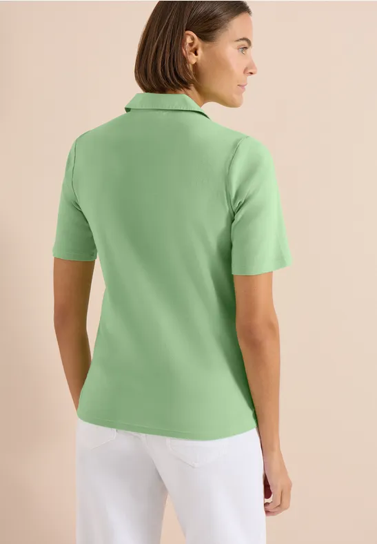 Rückenansicht einer Frau in hellgrünem Poloshirt und weißen Hosen vor hellem, neutralem Hintergrund.