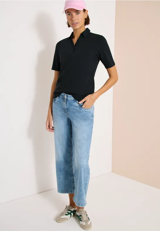 Person in schwarzem Poloshirt, hellblauen, weiten Cropped-Jeans und grün-weißen Sneakern vor zweifarbiger Wand.