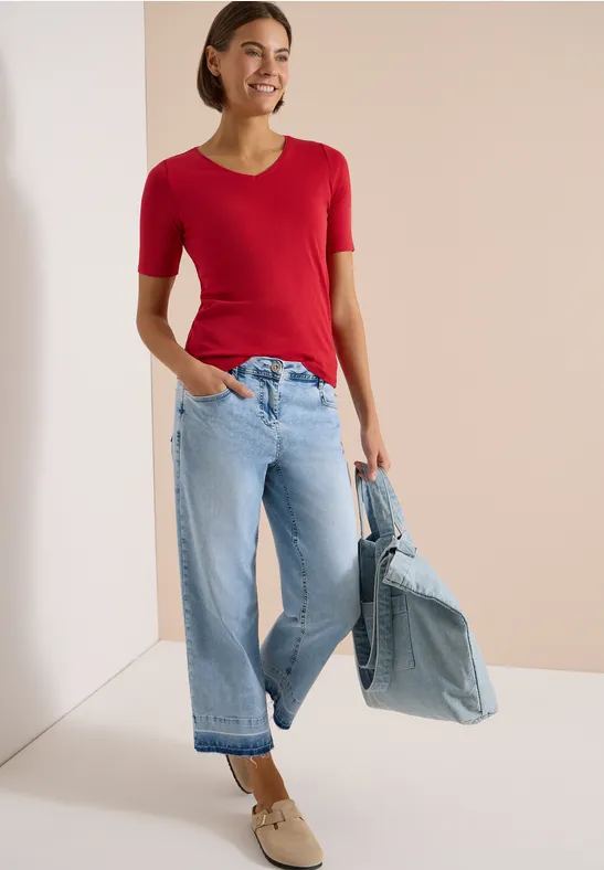 Frau trägt rotes Kurzarmshirt, weite hellblaue Jeans, hält lässig eine Jeansjacke, beige Schuhe.