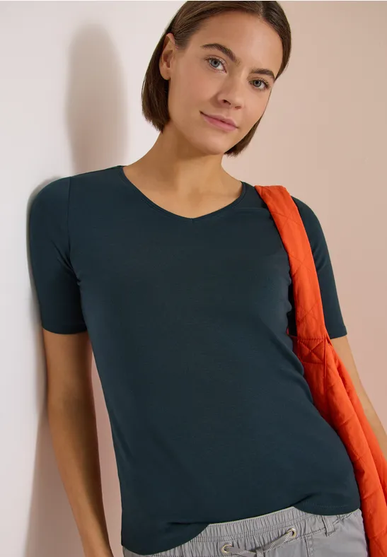 Jemand lehnt entspannt in einem dunkelblauen T-Shirt an einer Wand, mit einer orangenen Tasche über der Schulter.