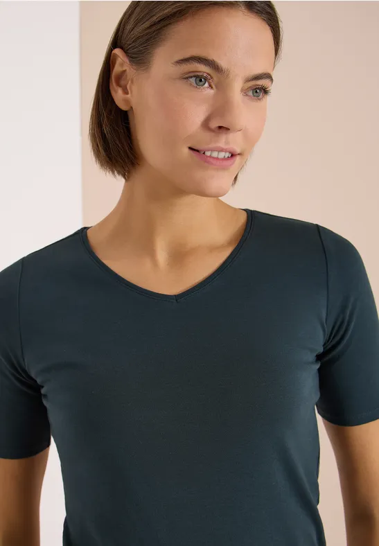 Eine Person trägt ein schlichtes, dunkelgrünes T-Shirt mit kurzen Ärmeln vor hellem Hintergrund.