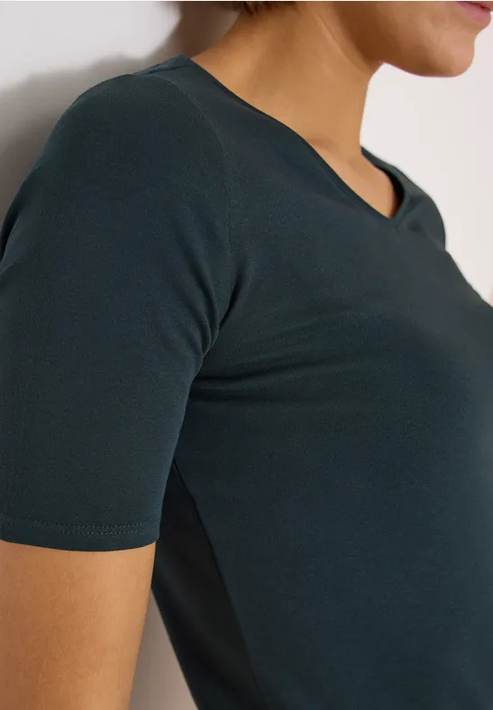 Seitliche Nahaufnahme einer Frau in einem dunkelgrünen, kurzärmligen V-Ausschnitt-Shirt.