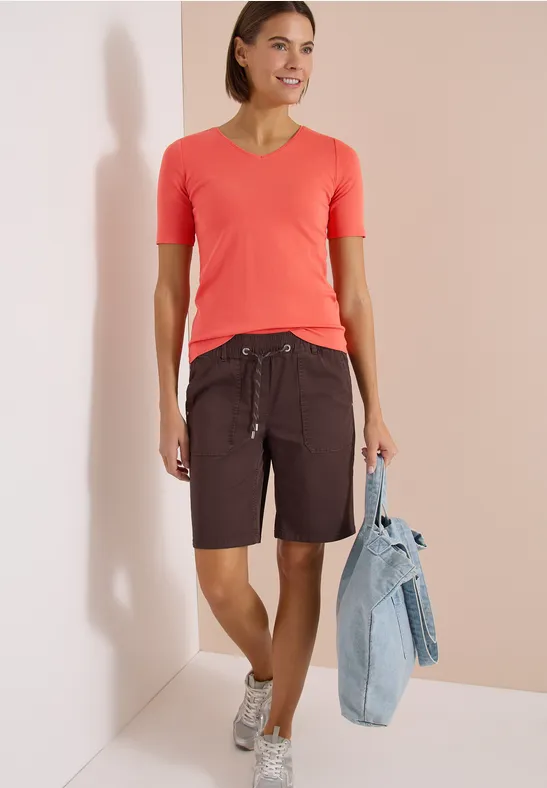 Frau in korallefarbenem V-Shirt, dunklen Bermudashorts mit Kordel, Sneakers und Jeansjacke.