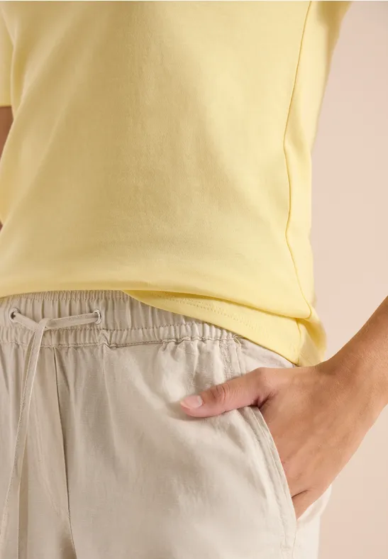 Nahaufnahme eines hellgelben T-Shirts und beiger Hose, Hand steckt in Hosentasche.