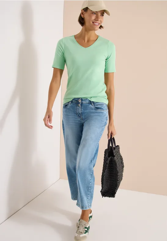 Frau in hellgrünem T-Shirt, blauen Jeans und Baseballcap trägt schwarze Tasche und weiße Sneaker.