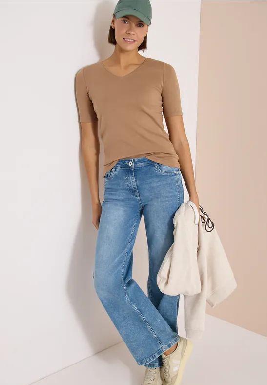 Vrouw leunt tegen muur, draagt beige V-hals shirt, blauwe jeans en houdt een lichte jas vast.