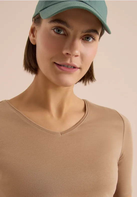 Persoon met groene pet en beige V-hals-shirt voor lichte achtergrond, gezicht onscherp.