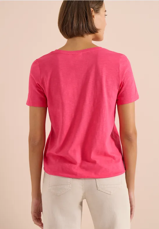 Een vrouw van achteren draagt een roze t-shirt en lichte broek voor een beige achtergrond.