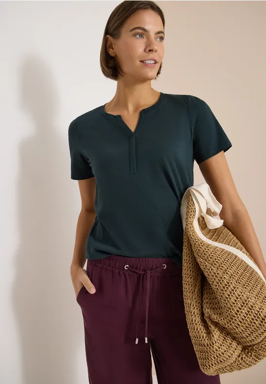 Frau in dunklem T-Shirt und weinroter Hose, trägt eine große, geflochtene Strohtasche.