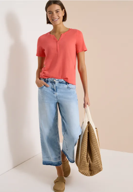 Frau in rotem T-Shirt und weiten Jeans, mit großer geflochtener Tasche lehnt an weißer Wand.
