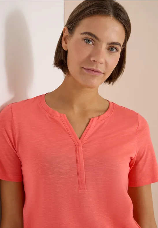 Eine Person trägt ein korallenfarbenes Kurzarmshirt mit Schlitzkragen vor hellem Hintergrund.