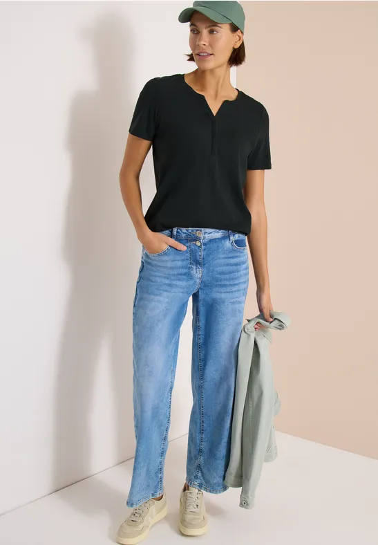 Persoon draagt jeans, zwart T-shirt en pet, houdt een jas vast, staat voor een lichte achtergrond.