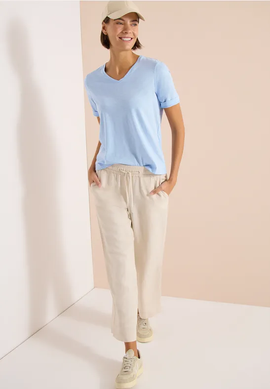 Person trägt hellblaues T-Shirt, bequeme Leinenhosen und Sneaker, steht entspannt in einem hellen Raum.