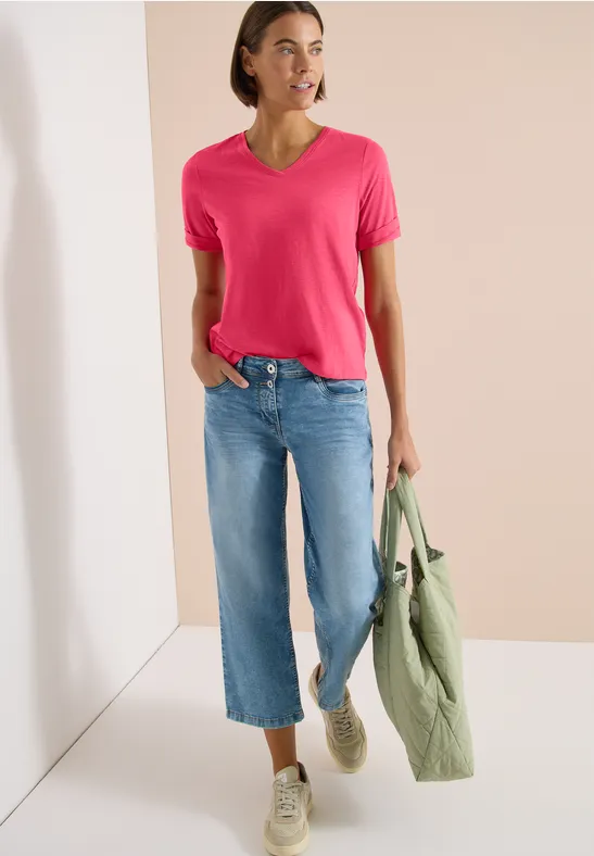 Model draagt roze V-hals T-shirt, wijde blauwe jeans en witte sneakers, met een groene jas in de hand.