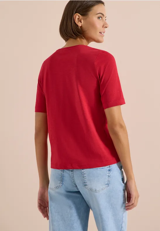Person von hinten mit rotem T-Shirt und hellblauen Jeans vor neutralem Hintergrund.