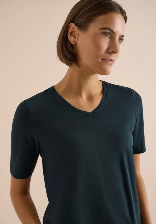Eine Person mit kurzem Haar trägt ein dunkelgrünes T-Shirt vor hellem Hintergrund.