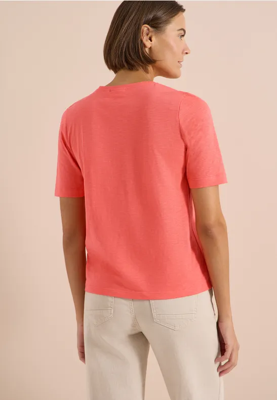 Frau von hinten in korallfarbenem T-Shirt und beige Hose vor hellem Hintergrund