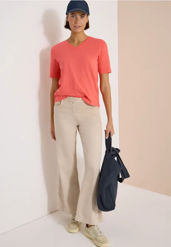 Person vor Wand in korallfarbenem T-Shirt, heller weiter Hose und Sneakers, hält Tasche.