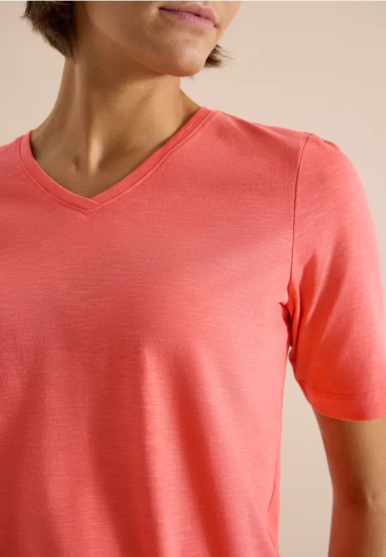 Person trägt ein korallenfarbenes V-Ausschnitt-Shirt vor neutralem Hintergrund.