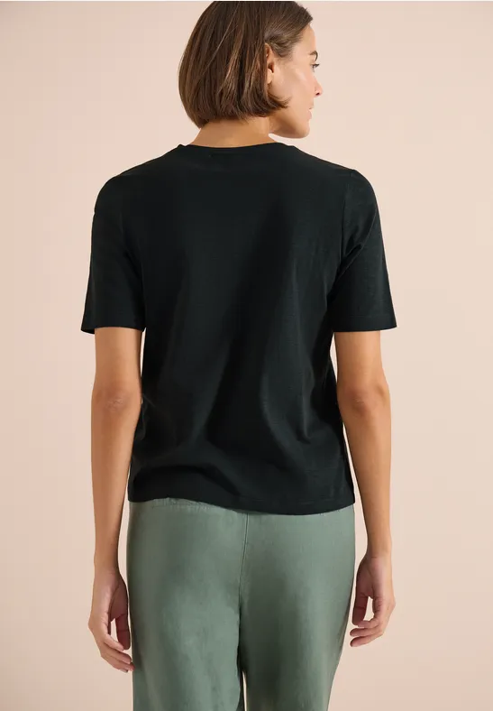 Frau von hinten in schwarzem T-Shirt und grünen Hosen vor neutralem Hintergrund