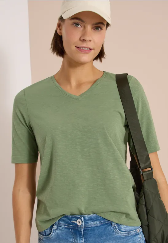 Frau in grünem V-Ausschnitt-Shirt und Basecap, mit Umhängetasche vor hellem Hintergrund.