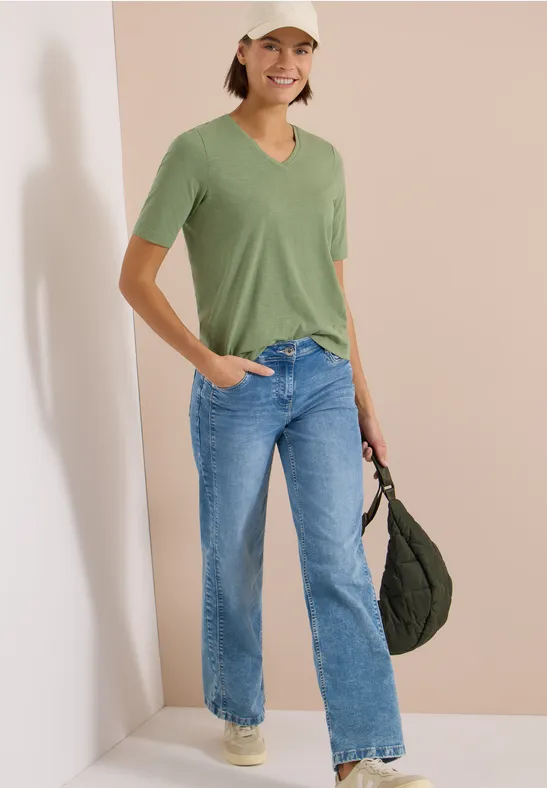 Frau trägt grünes T-Shirt, blaue Jeans, weiße Sneaker, Baseballcap und hält eine dunkelgrüne Umhängetasche.