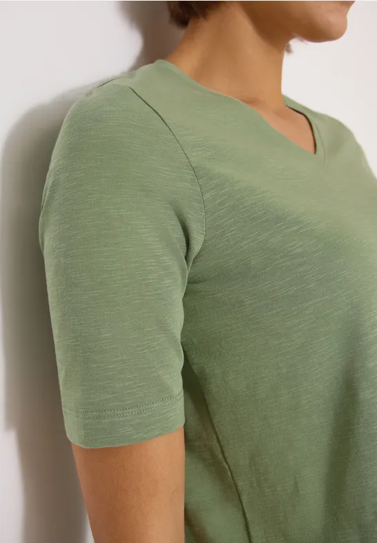 Frau in hellgrünem V-Ausschnittshirt mit kurzen Ärmeln, seitliche Ansicht, oberer Oberkörper sichtbar.