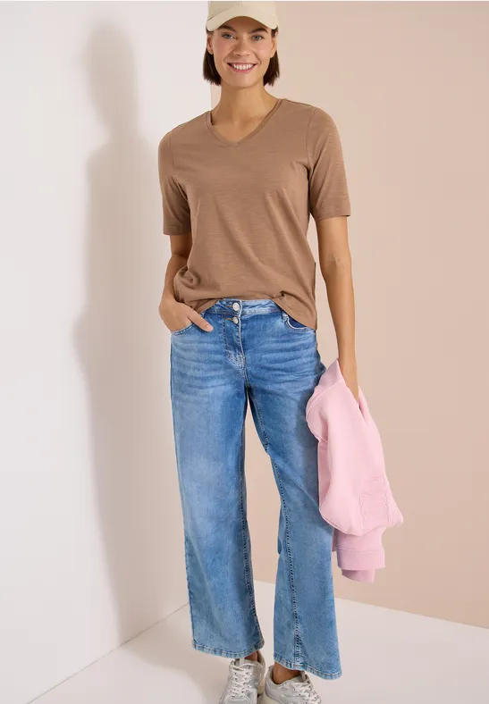 Vrouw in bruin T-shirt en blauwe jeans met roze trui voor een lichte muur.