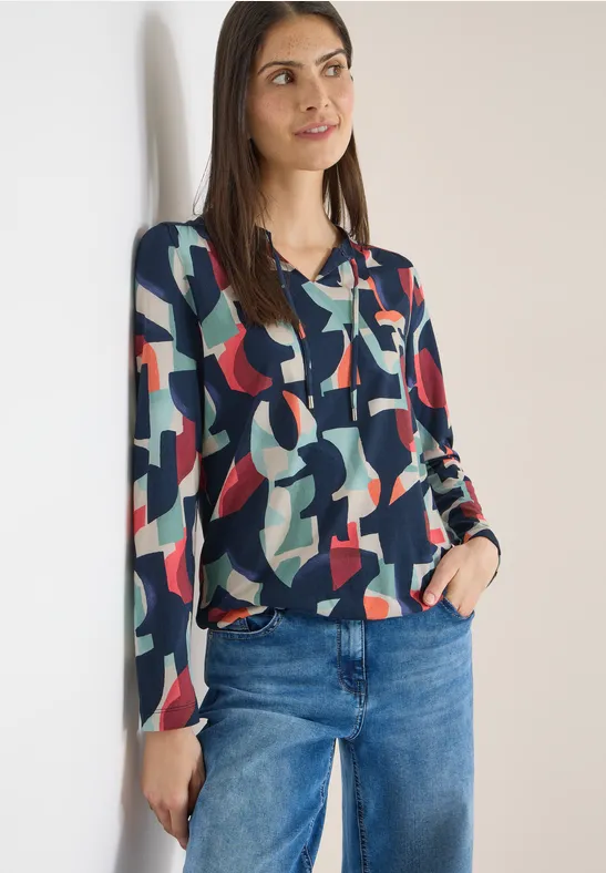 Eine Person trägt eine bunte Bluse mit geometrischem Muster und blaue Jeans.